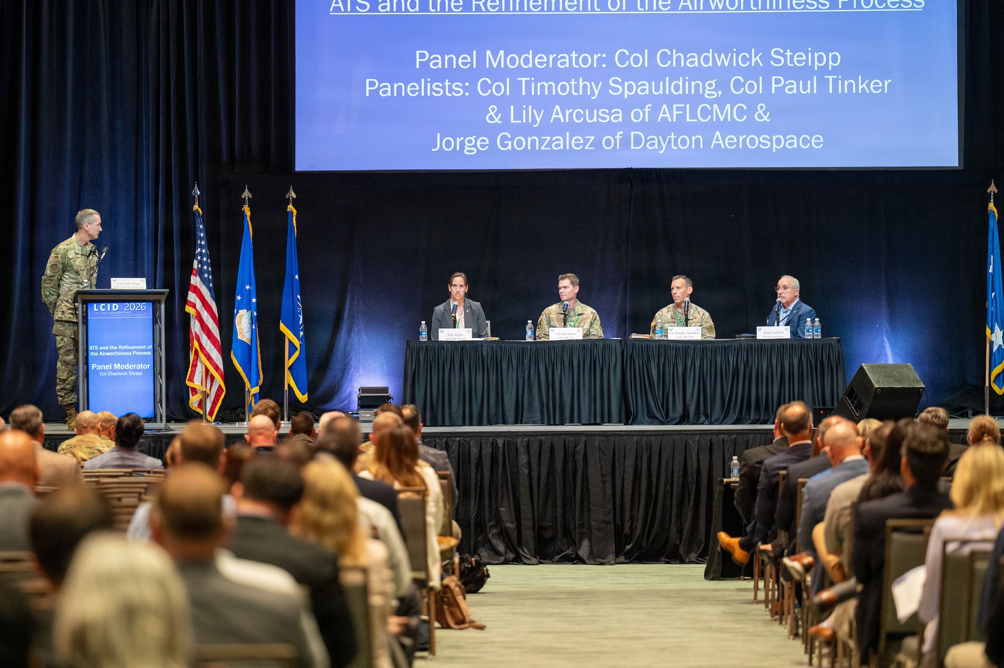 A large conference hall, with a panel of four people seated on the stage, and a moderator at a podium.