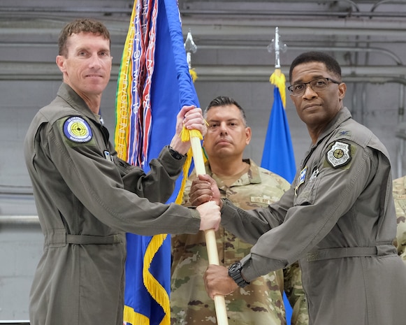 photo of three Airmen standing on a stage, two Airmen are holding a unit flag in between them