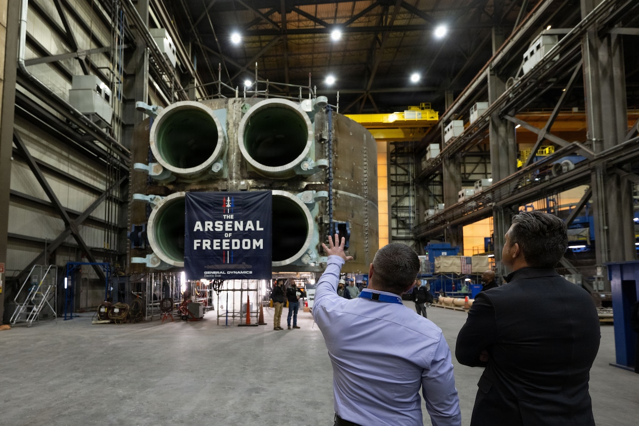 Two men stand near each other and look at a large piece of military hardware inside a large building. A sign hangs off the equipment that says, "The Arsenal of Freedom."