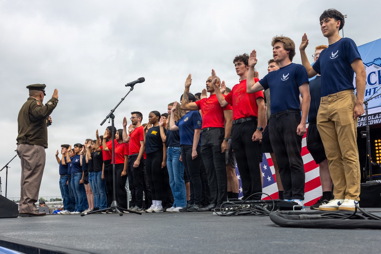 More than a dozen young people stand together on a stage and raise their right hands as a man in a formal military uniform stands in front of them with his own hand raised.