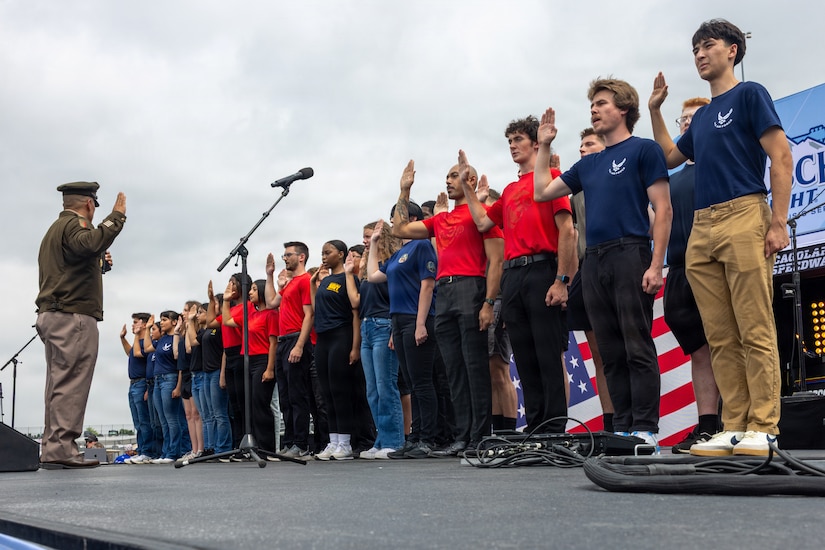 More than a dozen young people stand together on a stage and raise their right hands as a man in a formal military uniform stands in front of them with his own hand raised.
