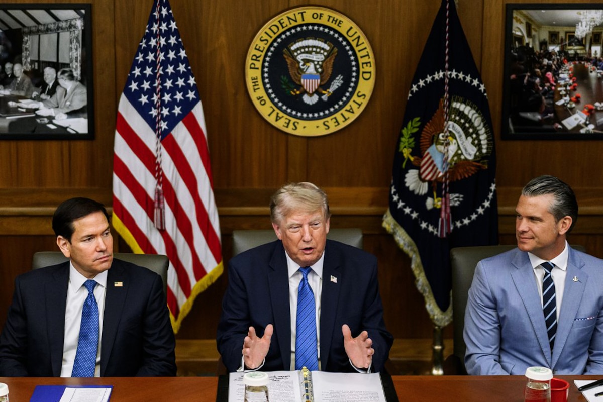 Three men in business attire sit next to each other at a table; behind them are an American flag and another flag.
