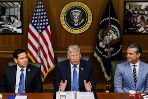 Three men in business attire sit next to each other at a table; behind them are an American flag and another flag.