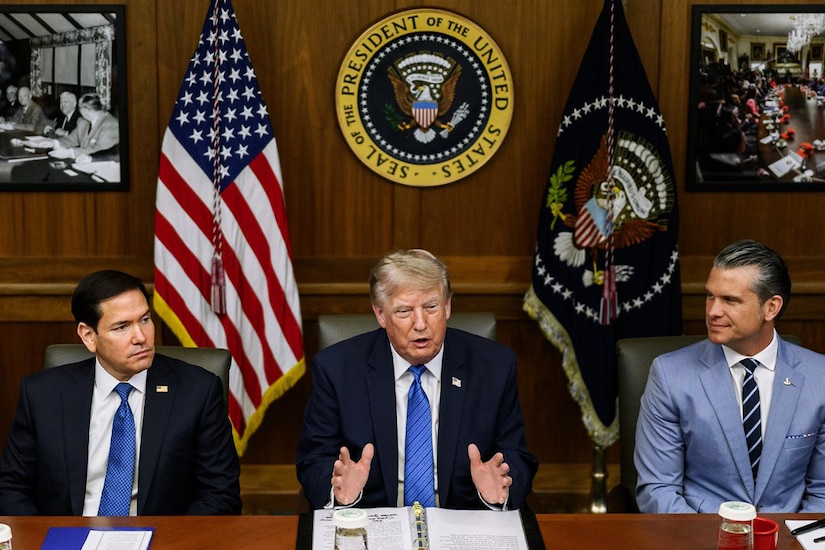 Three men in business attire sit next to each other at a table; behind them are an American flag and another flag.