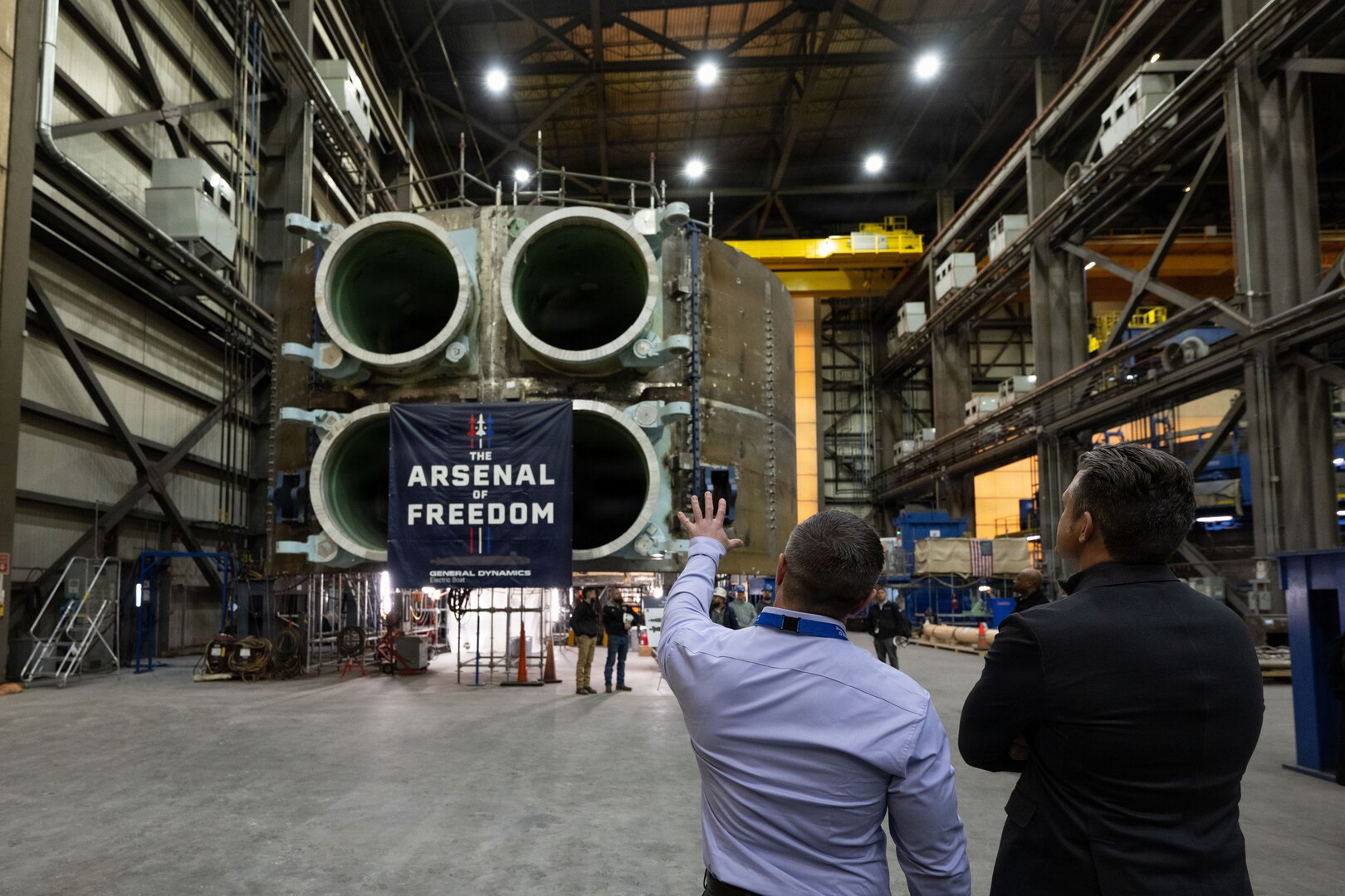 Two men stand near each other and look at a large piece of military hardware inside a large building. A sign hangs off the equipment that says, "The Arsenal of Freedom."