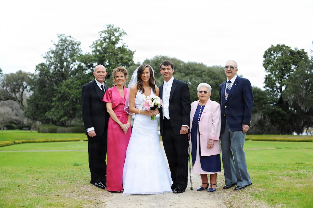 A wedding group stands outdoors on a dirt path with a green lawn and trees in the background, featuring a bride and groom in the center flanked by two older couples, including the bride's grandparents, Edward Hoenshel and his wife.