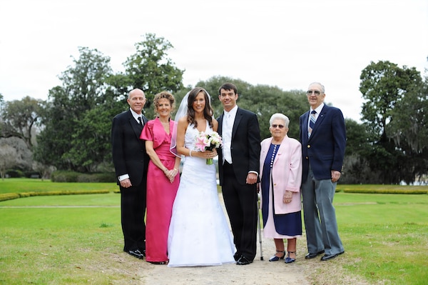 A wedding group stands outdoors on a dirt path with a green lawn and trees in the background, featuring a bride and groom in the center flanked by two older couples, including the bride's grandparents, Edward Hoenshel and his wife.