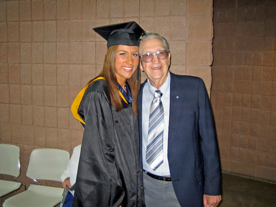 A smiling Ashlee Landreth in a black graduation gown and cap stands alongside her grandfather, Edward Hoenshel, who wears a blue suit jacket and a striped tie against a brick wall background.