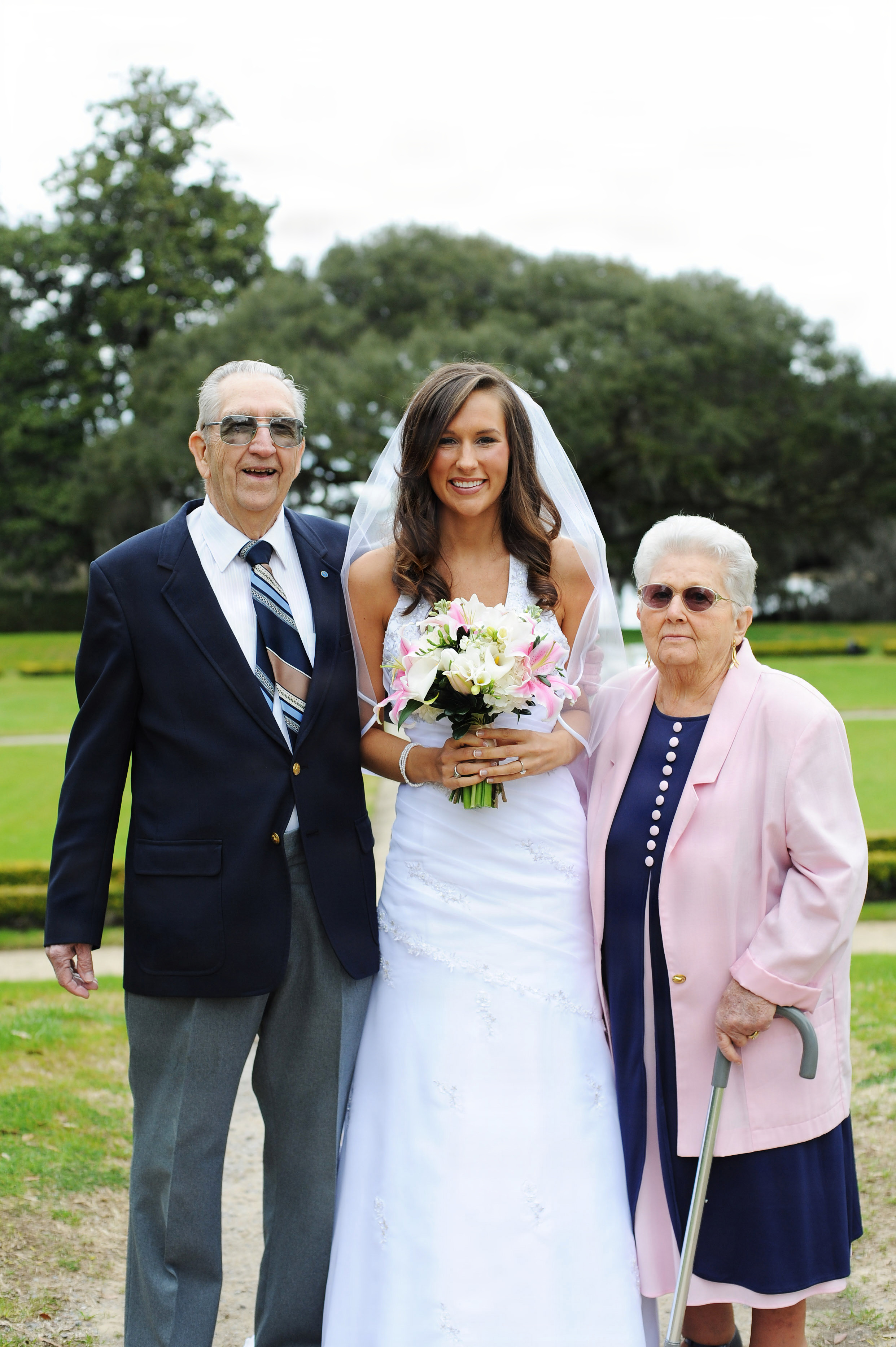 A smiling bride in a white gown and veil stands outdoors between her grandparents, Edward Hoenshel and his wife, holding a bouquet of pink and white lilies.
