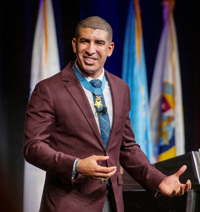 A man, wearing a business suit and the Medal of Honor, gestures with his hands as he speaks while standing on a stage indoors. There are a variety of flags behind him.