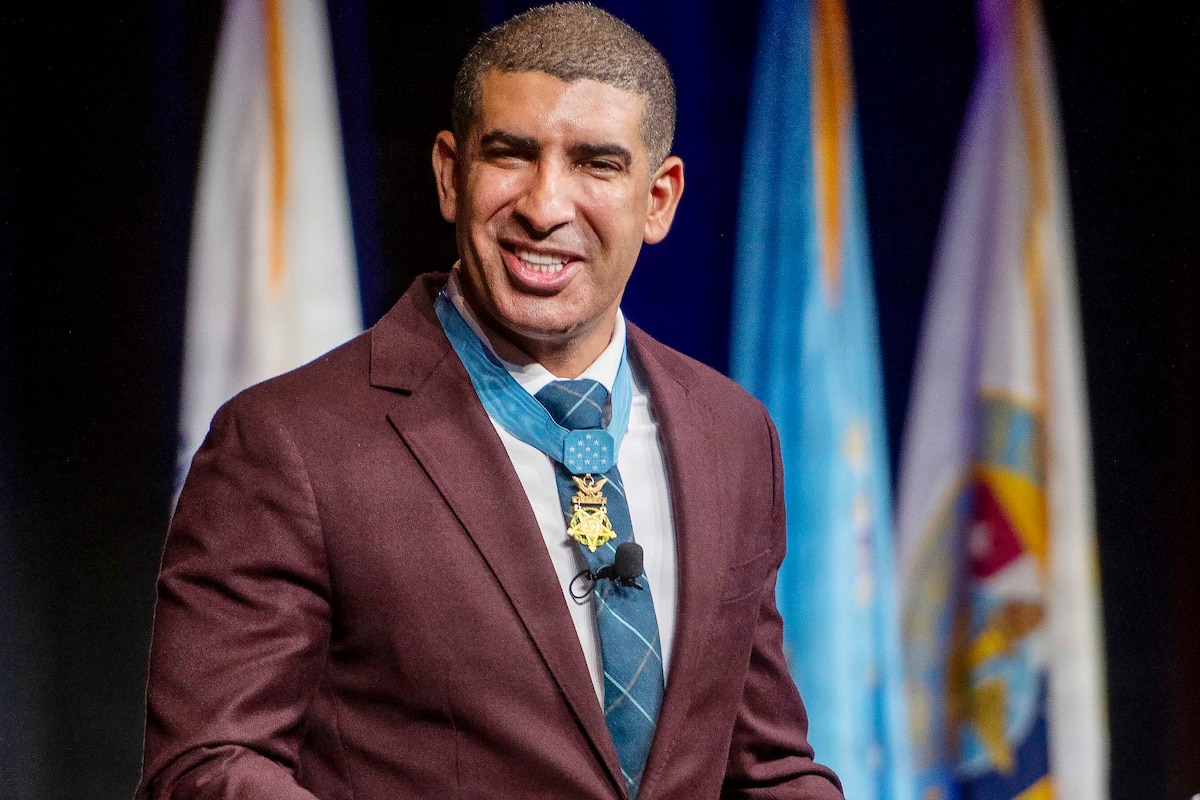 A man, wearing a business suit and the Medal of Honor, gestures with his hands as he speaks while standing on a stage indoors. There are a variety of flags behind him.
