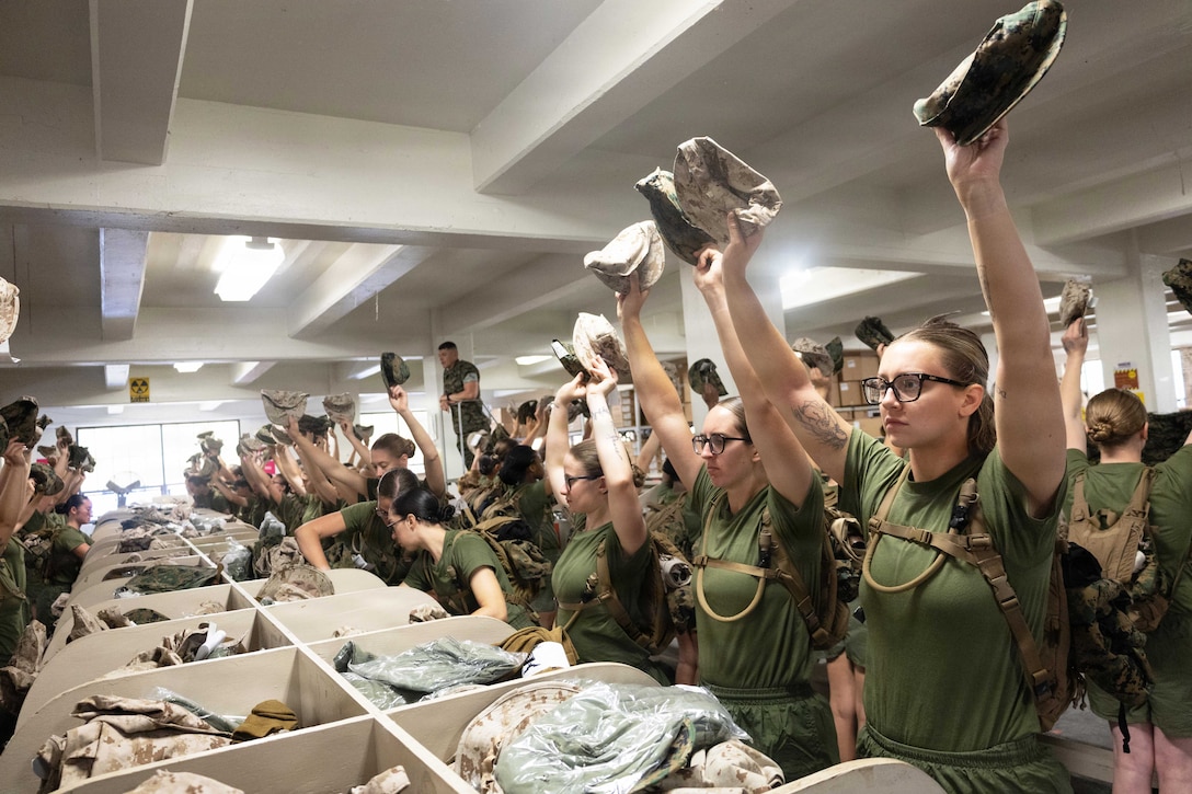 Marine Corps officer candidates in green T-shirts and shorts raise their hands while holding hats in front of cubbies filled with uniforms.