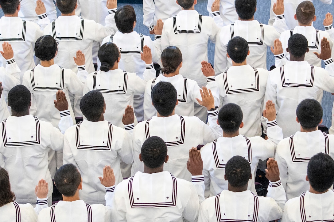 Dozens of Navy recruits in ceremonial dress raise their right hands as seen from behind.