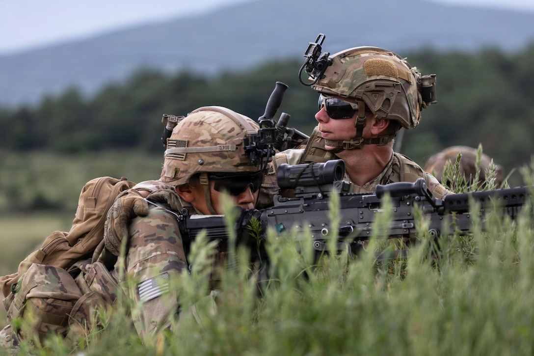 A soldier in tactical gear kneels behind grass while looking through the scope of a weapon as a fellow soldier keeps watch.