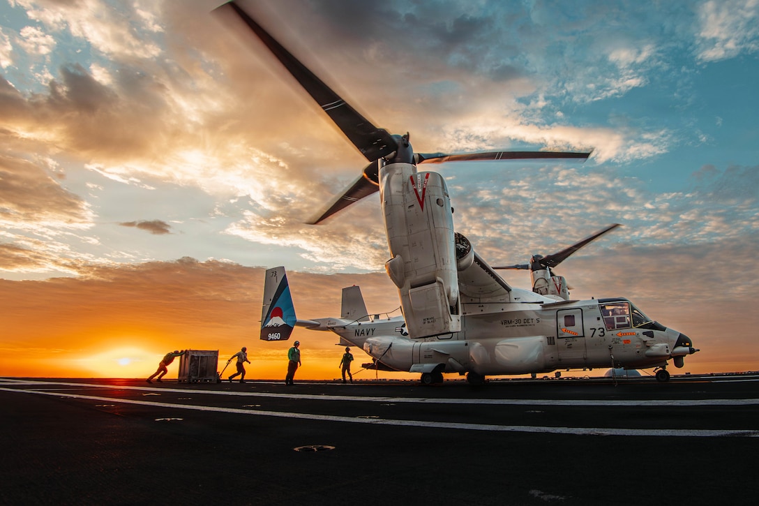 Two sailors push and pull cargo toward an aircraft on a ship's flight deck as two others stand nearby, with an orangish, sunlit sky over the horizon in the background.