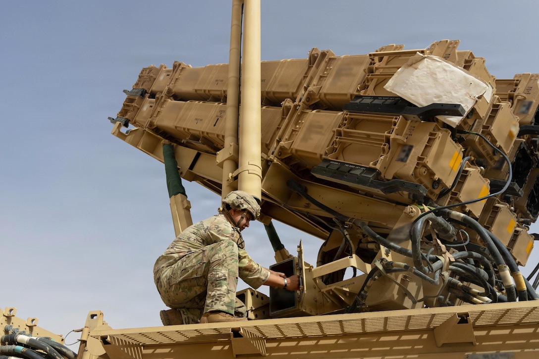 A soldier kneels to work on a missile against a blue sky.