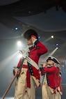 Army Soldiers in red, Revolutionary War-era uniforms are marching in formation during an indoor event with bright spotlights shining from the ceiling. The soldier in the front has a large, furry hat and is saluting, while soldiers behind are performing on musical instruments.