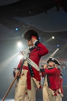 Army Soldiers in red, Revolutionary War-era uniforms are marching in formation during an indoor event with bright spotlights shining from the ceiling. The soldier in the front has a large, furry hat and is saluting, while soldiers behind are performing on musical instruments.