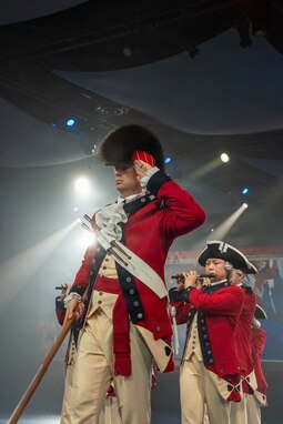 Army Soldiers in red, Revolutionary War-era uniforms are marching in formation during an indoor event with bright spotlights shining from the ceiling. The soldier in the front has a large, furry hat and is saluting, while soldiers behind are performing on musical instruments.