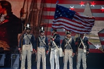 Five Army Soldiers wearing 1800s-era period uniforms stand in a row while the middle soldier waves a historic U.S. flag during an indoor show.