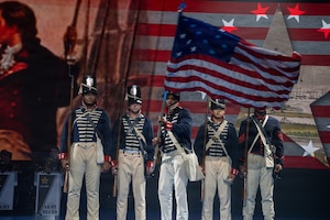 Five Army Soldiers wearing 1800s-era period uniforms stand in a row while the middle soldier waves a historic U.S. flag during an indoor show.