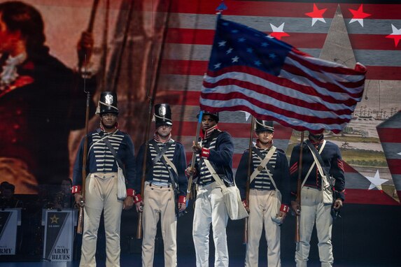 Five Army Soldiers wearing 1800s-era period uniforms stand in a row while the middle soldier waves a historic U.S. flag during an indoor show.
