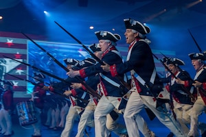 Army Soldiers in Revolutionary War-era uniforms run while yelling and carrying period rifles with bayonets affixed during an indoor show.