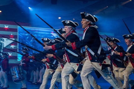 Army Soldiers in Revolutionary War-era uniforms run while yelling and carrying period rifles with bayonets affixed during an indoor show.