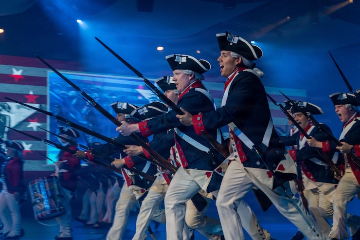 The U.S. Army Old Guard Fife and Drum Corps, wearing red Revolutionary War-era uniforms, is standing in formation during a Twilight Tattoo performance in Conmy Hall.