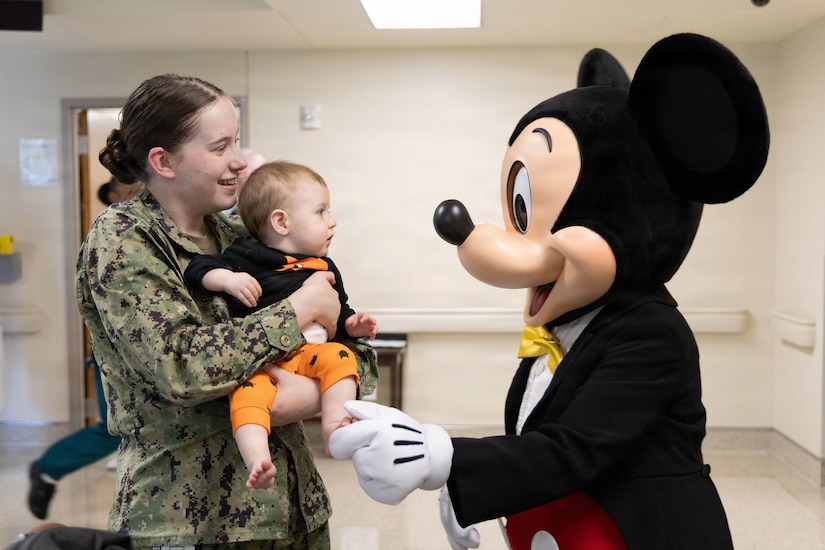 A woman in a camouflage military uniform holds a baby in a hospital hallway and greets a person in a Mickey Mouse costume.