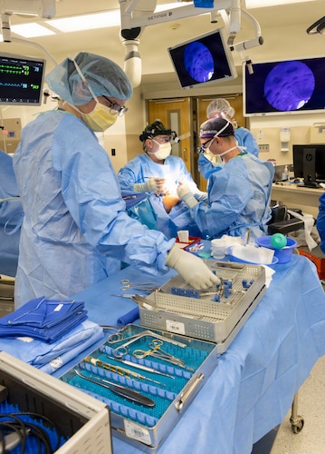 Four people in surgical gowns operate on a patient in a hospital room surrounded by medical equipment and screens.
