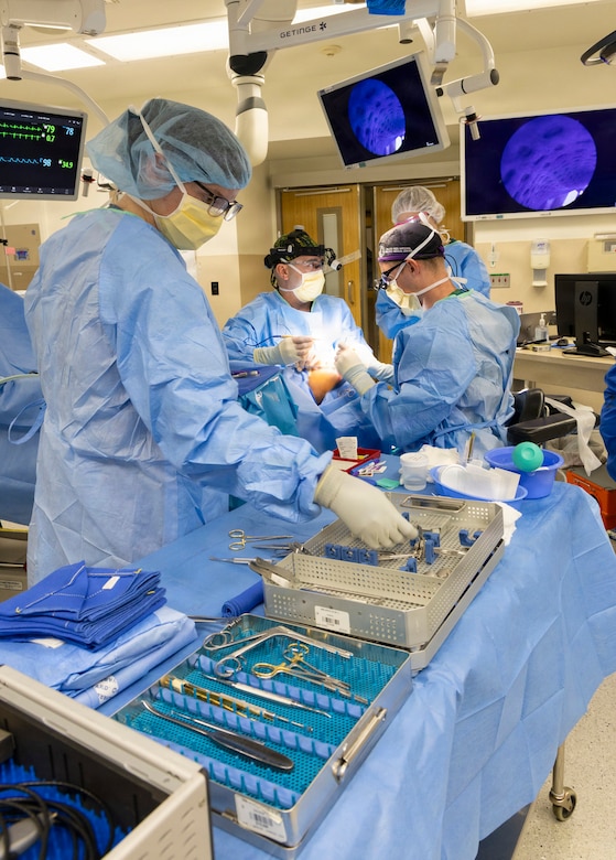 Four people in surgical gowns operate on a patient in a hospital room surrounded by medical equipment and screens.