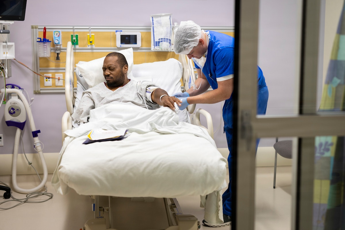 A man in a hospital gown lies in a hospital bed while another man in medical scrubs examines him.