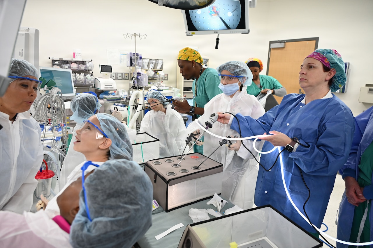 Nearly a dozen adults and kids, all in surgical gowns and hair nets, stand in a hospital room and look at medical equipment.