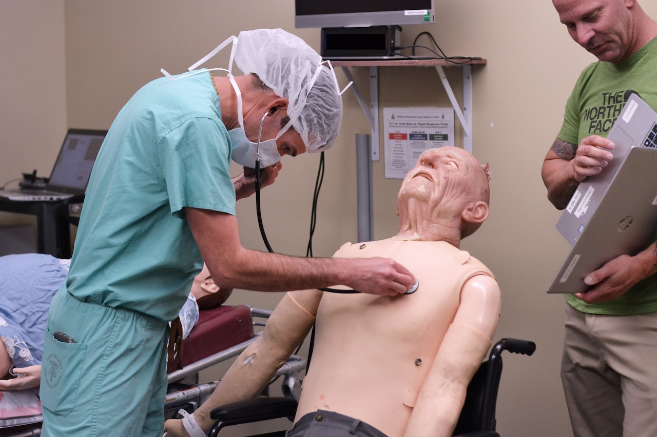 A man in medical scrubs, hair net and a face mask uses a stethoscope on a medical dummy while another man in casual attire observes.