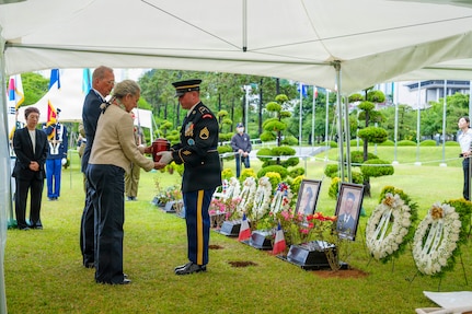 Staff at the United Nations Memorial Cemetery Korea present a flag‑draped urn containing the remains of French Korean War veteran Jacques Grisolet to his family during an interment ceremony in Busan, South Korea, on May 27, 2026. Family members stand solemnly as cemetery officials complete the handoff, honoring both Grisolet and fellow veteran André Datcharry. The scene takes place outdoors among manicured grounds and memorial markers, reflecting the ceremony’s recognition of France’s sacrifice during the Korean War and its ongoing support to the United Nations Command.