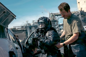 A service member wearing a brown t-shirt helps another service member adjust his black protective full body suit and breathing aparatus.