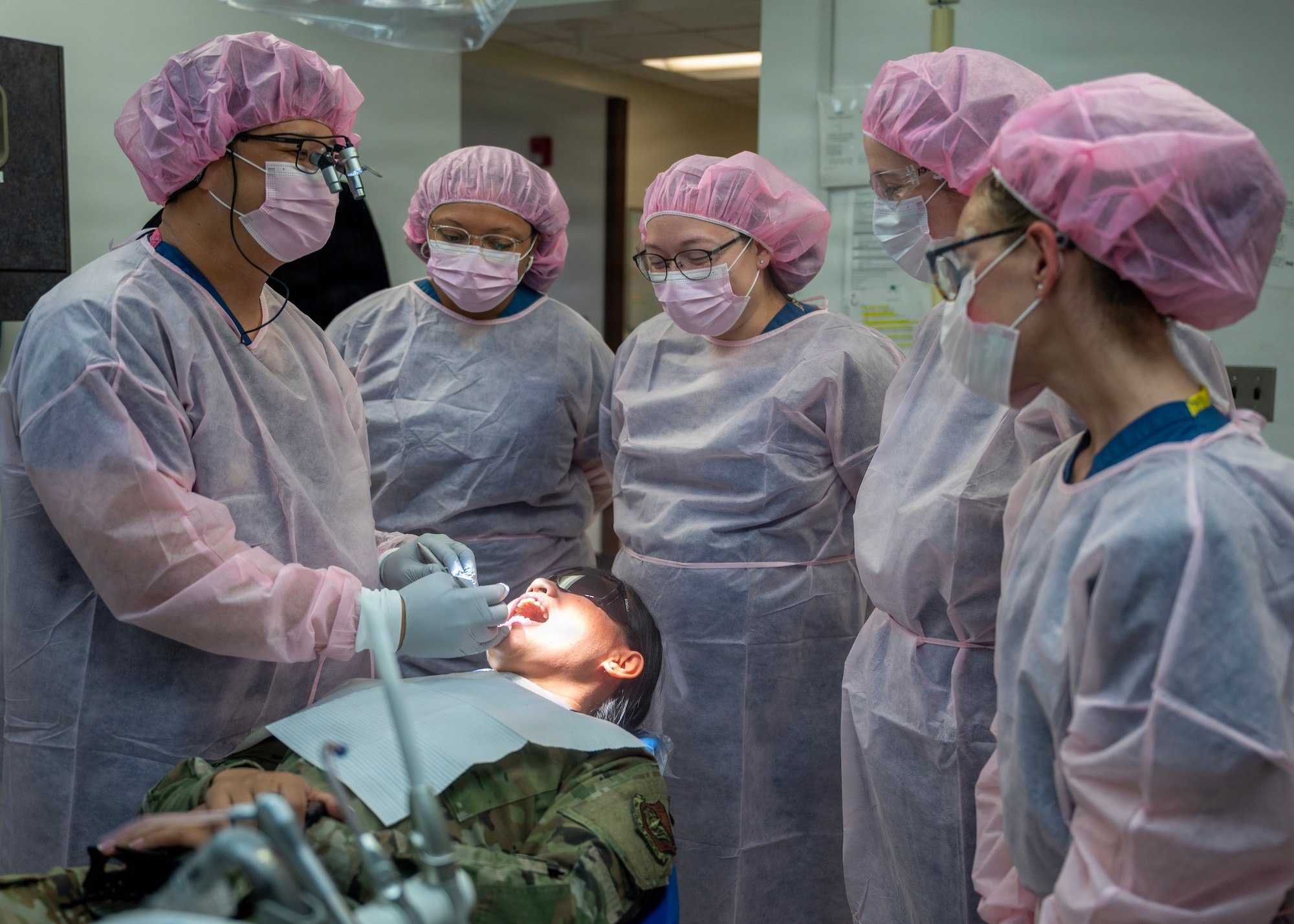 Five people in scrubs and hairnets observe a dental exam of a uniformed person. The uniformed person is lying down with her mouth open while wearing protective eye coverings and a dental bib.