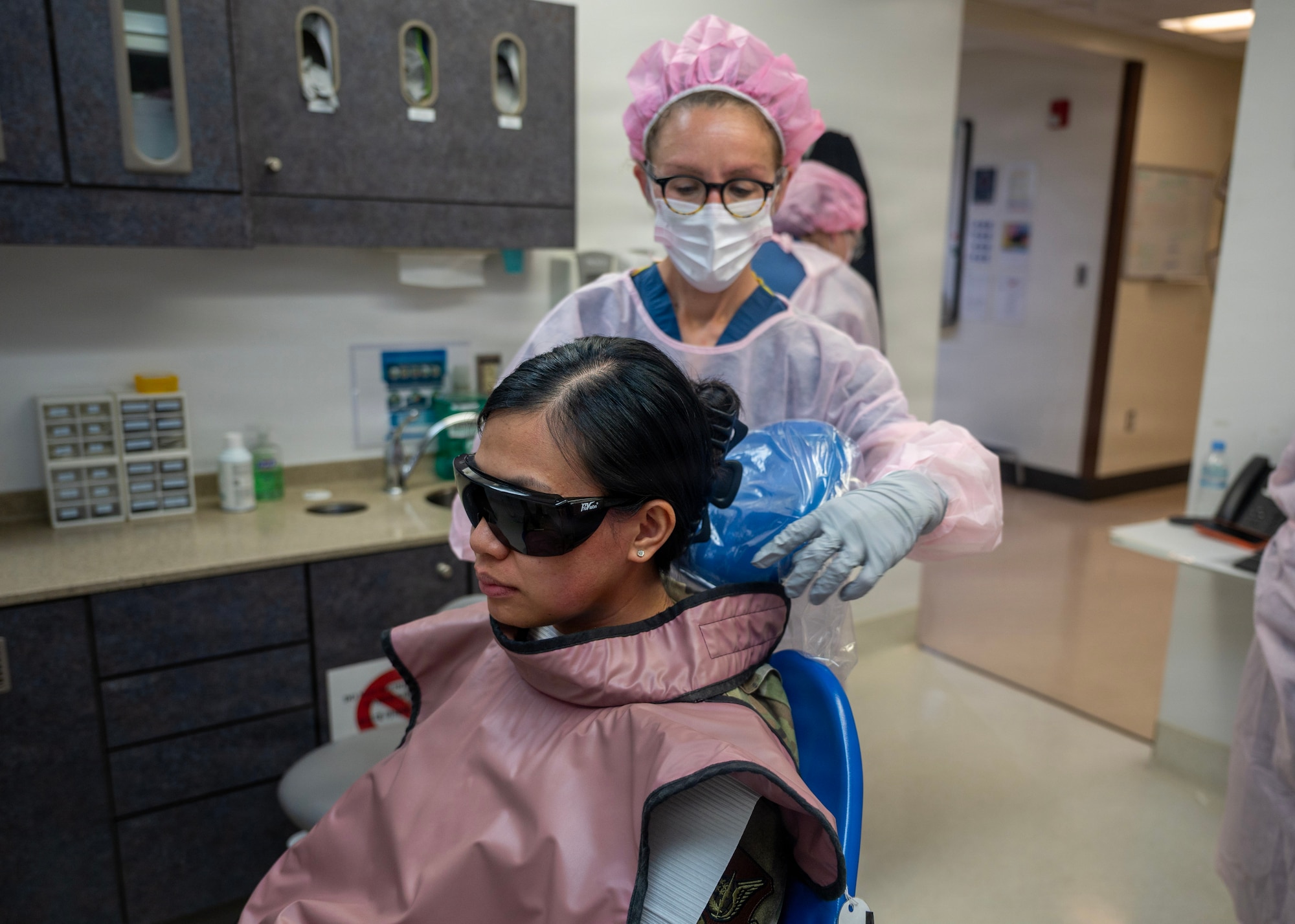 A person in scrubs and a pink hairnet drapes a cape over a woman wearing protective eyewear