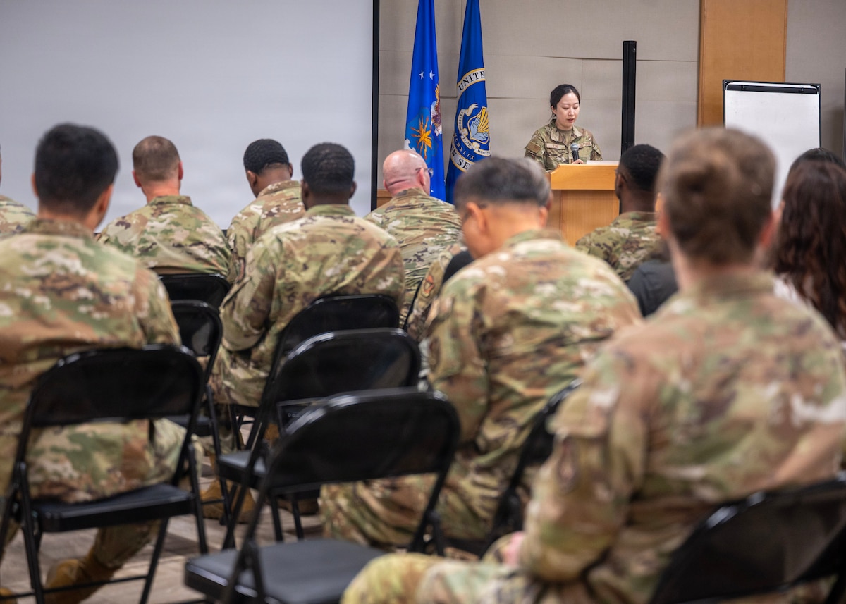 A group of military personnel are seated facing a podium and projector screen