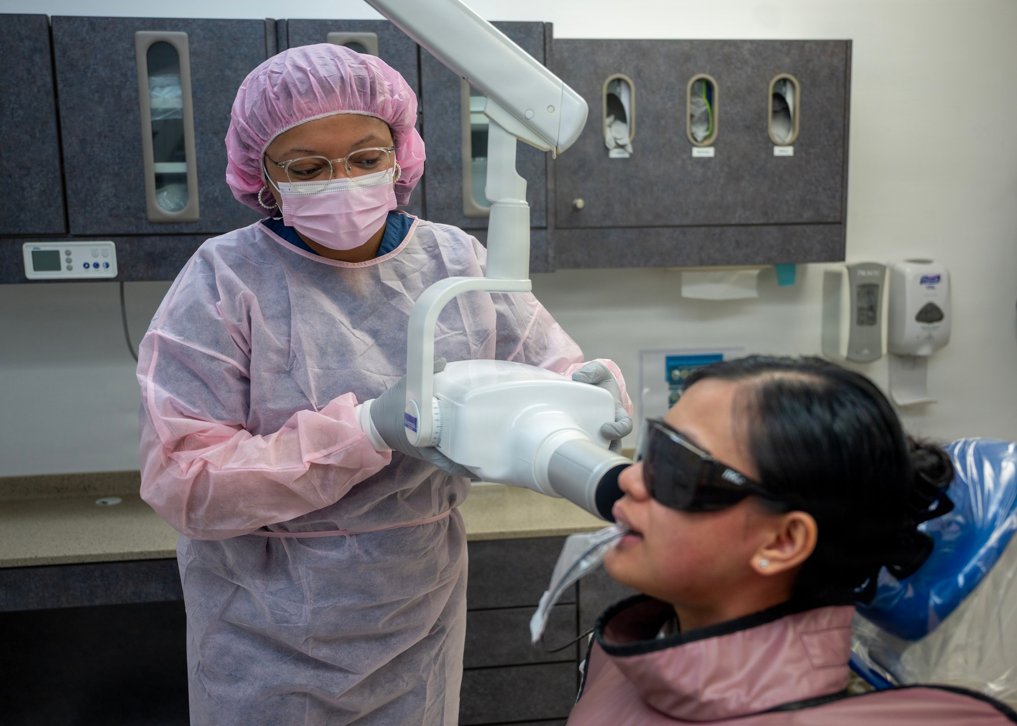 Woman wearing a pink hairnet and scrubs is seen giving a woman wearing protective eyewear a dental x-ray