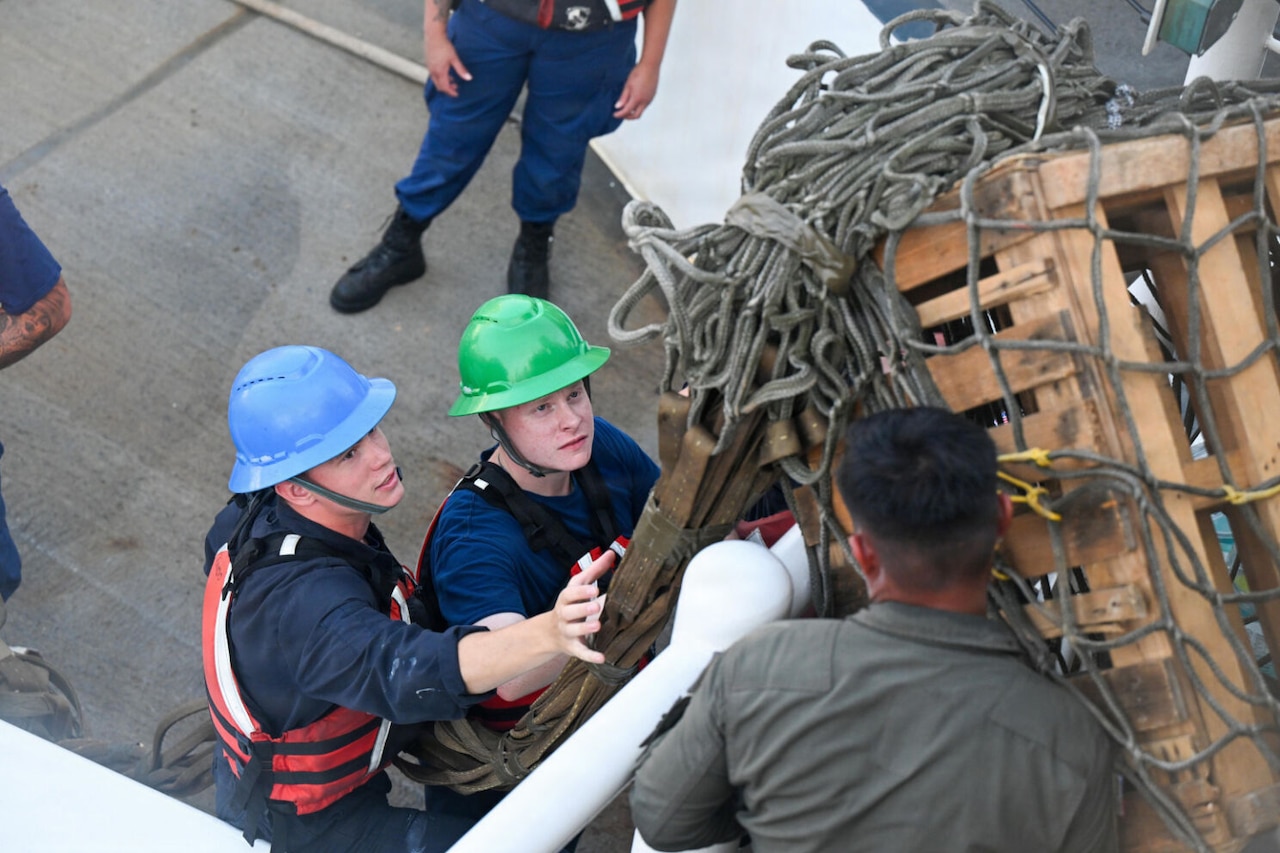 Two men wearing hard hats and life jackets stand on a ship and wait for another man to pass a wooden crate with ropes around it to them.
