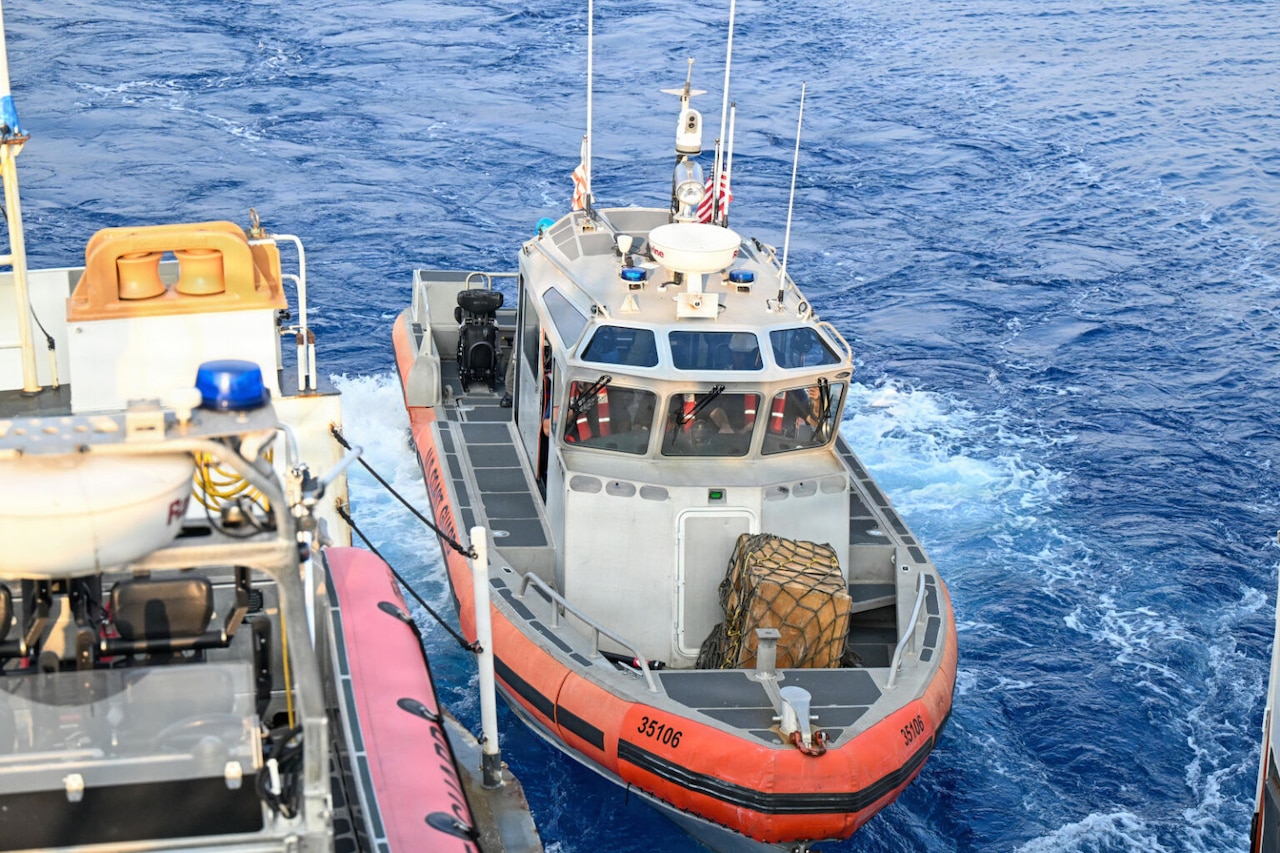 A small boat is tied to a large ship sailing in the ocean; three people wearing life jackets are in the cockpit of the boat with a package on the bow.