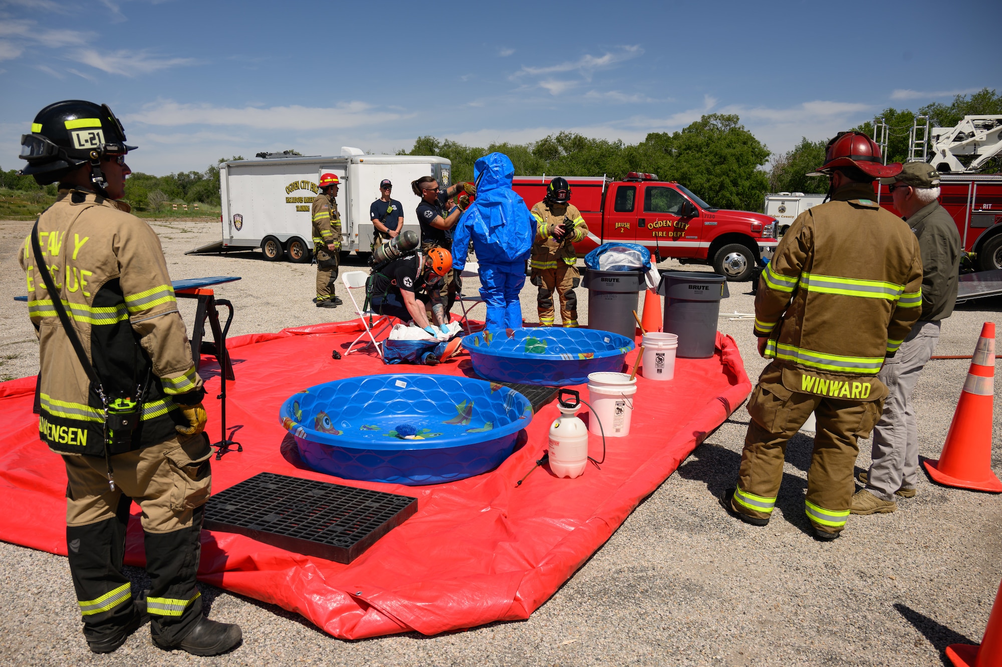 Firefighters observe as personnel in decontamination suits undergo cleaning procedures over two small pools on a red tarp. Buckets and sprayers are arranged nearby, with an emergency response vehicle parked in the background.