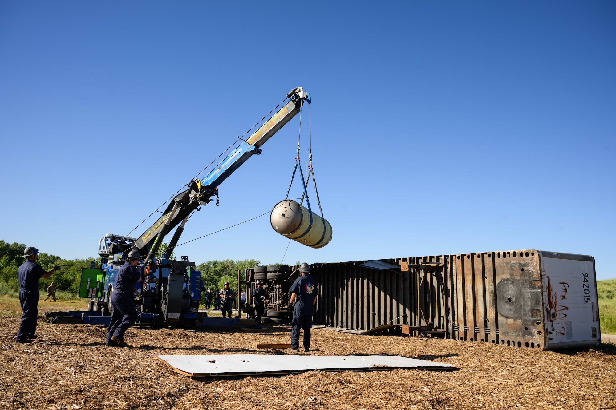 A crane hoists a simulated Minuteman III component over an overturned semi-trailer, with responders in blue coveralls and hard hats scattered throughout the scene.