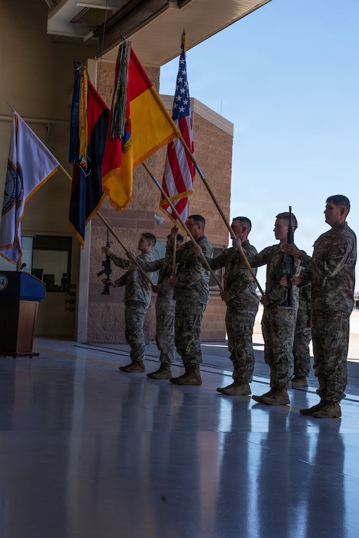 Six men in camouflage military uniforms stand in formation in an aircraft hangar. Three of the men are holding up various flags while one is holding a flag forward, away from himself.