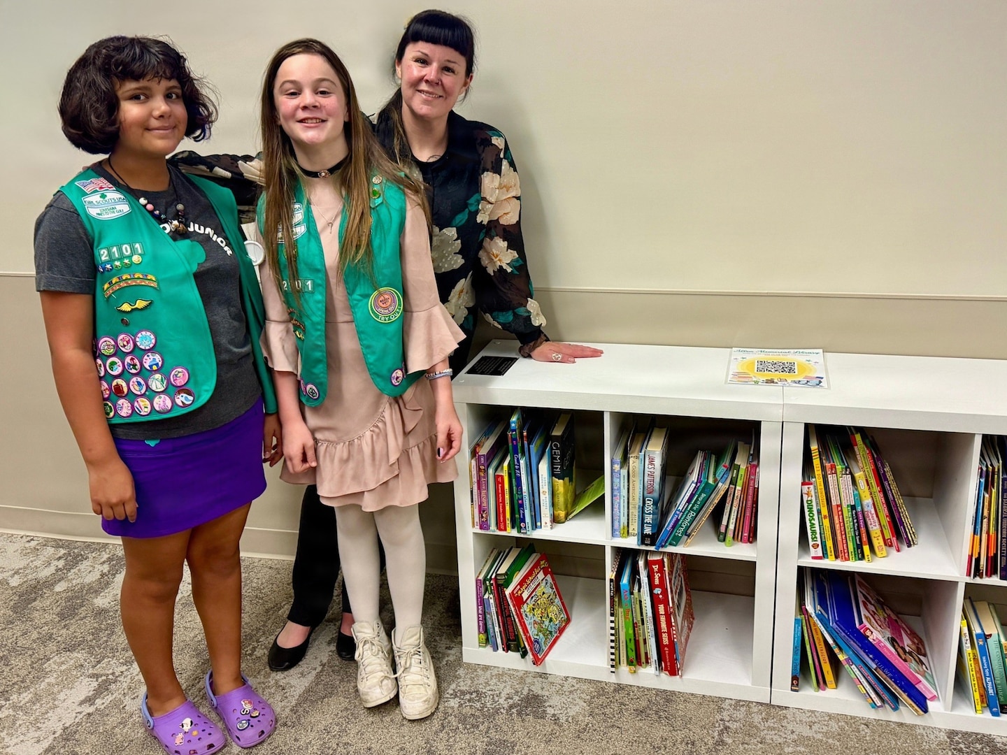 Two Girl Scouts pose with their Troop Leader in front of a shelf of donated books.