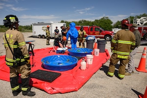 Firefighters observe as personnel in decontamination suits undergo cleaning procedures over two small pools on a red tarp. Buckets and sprayers are arranged nearby, with an emergency response vehicle parked in the background.