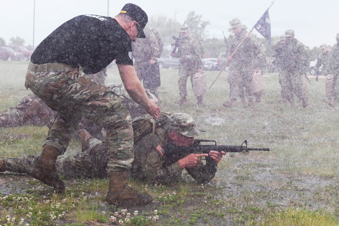 Heavy rain falls as a soldier directs an officer candidate lying on a muddy field aims a weapon as fellow candidates march in the background.