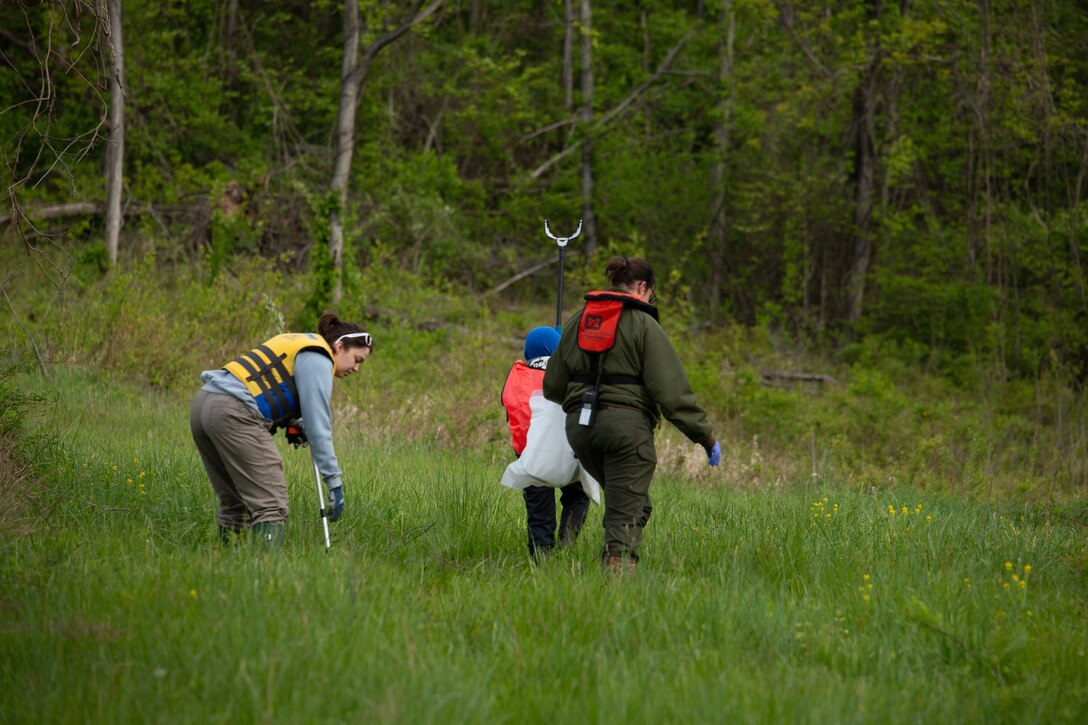 Two adults and a child, all wearing life jackets, walk down a hill with trash bags. The individual on the left leans over to pick up something from the grass.